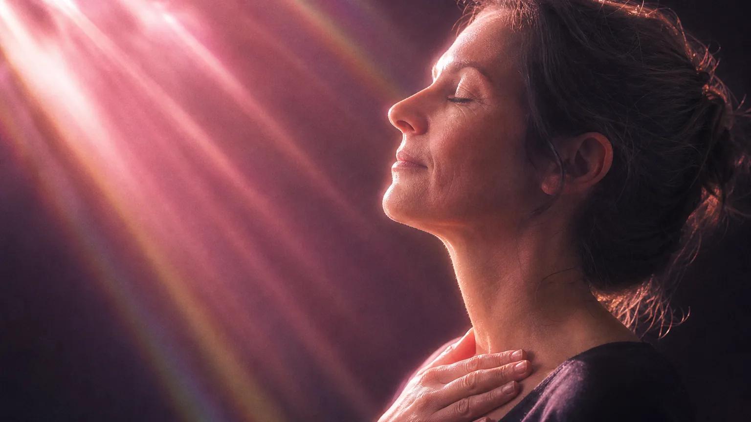 A woman in her 40s in profile, eyes closed, hand at her collarbone, in soft rose-pink light.